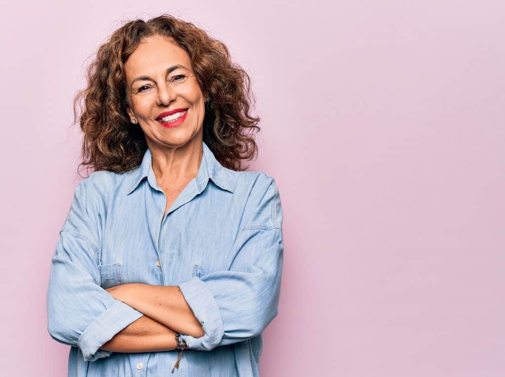 AdobeStock 354110985 - Viera Dental Arts | Dentist in Melbourne FL Middle age beautiful woman wearing casual denim shirt standing over pink background happy face smiling with crossed arms looking at the camera. Positive person.